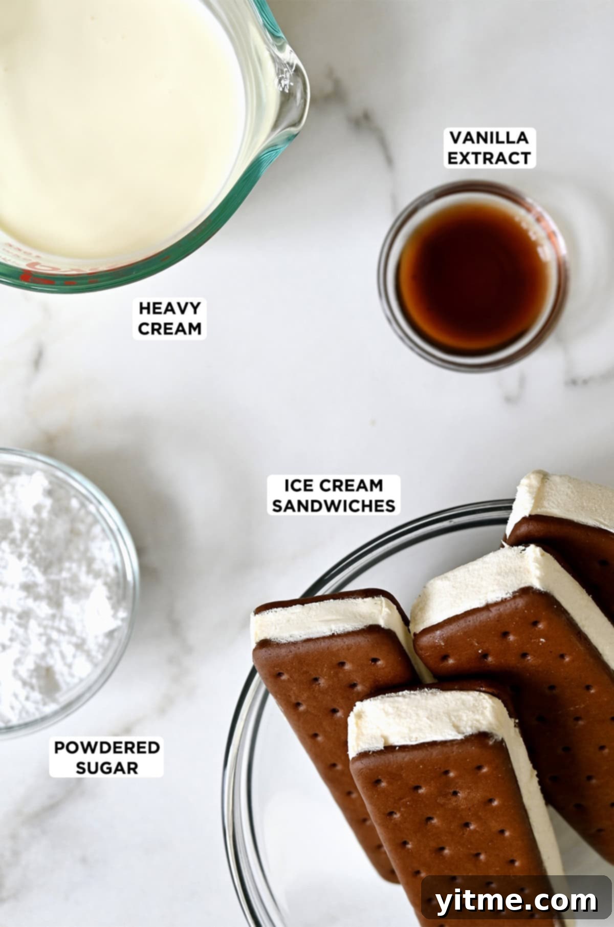Ice cream sandwiches in a glass bowl next to bowls containing powdered sugar, heavy cream and vanilla extract.