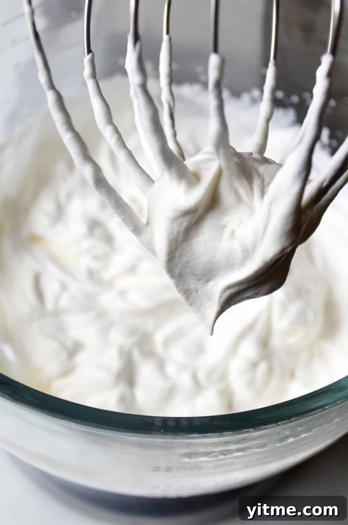 Stiff whipped cream peaks on a beater above a bowl with whipped cream.