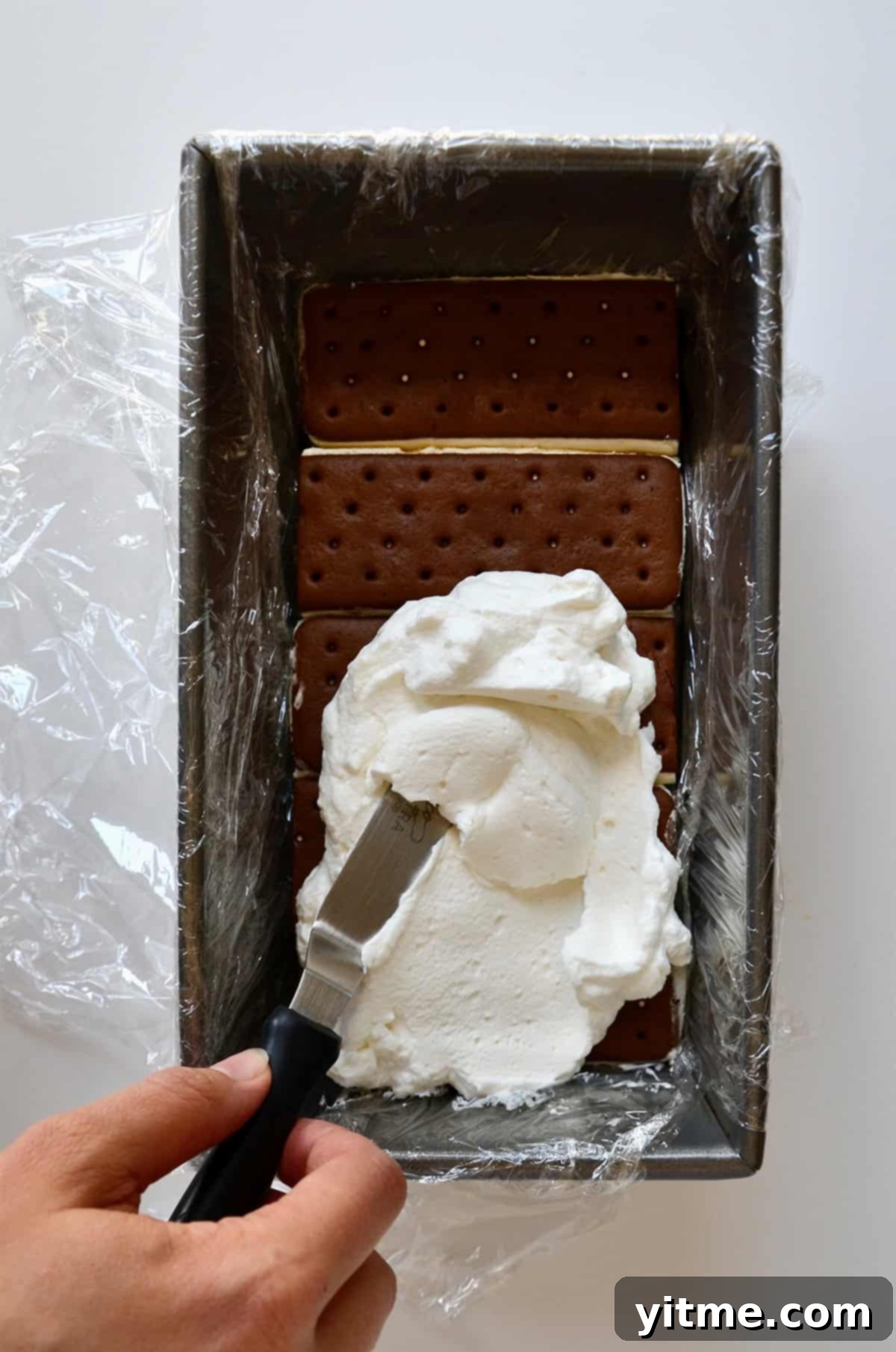 Whipped cream being spread with an offset spatula atop ice cream sandwiches in a loaf pan lined with plastic wrap.