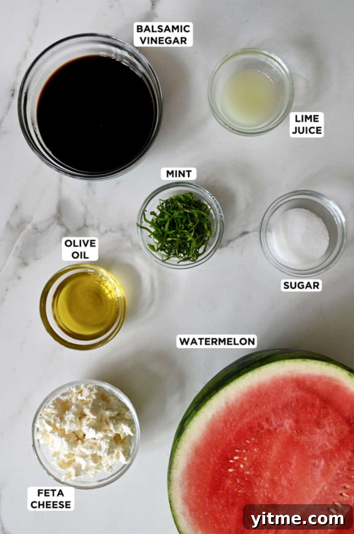 A large watermelon sliced in half next to small bowls containing feta cheese, olive oil, mint, sugar, lime juice, and balsamic syrup