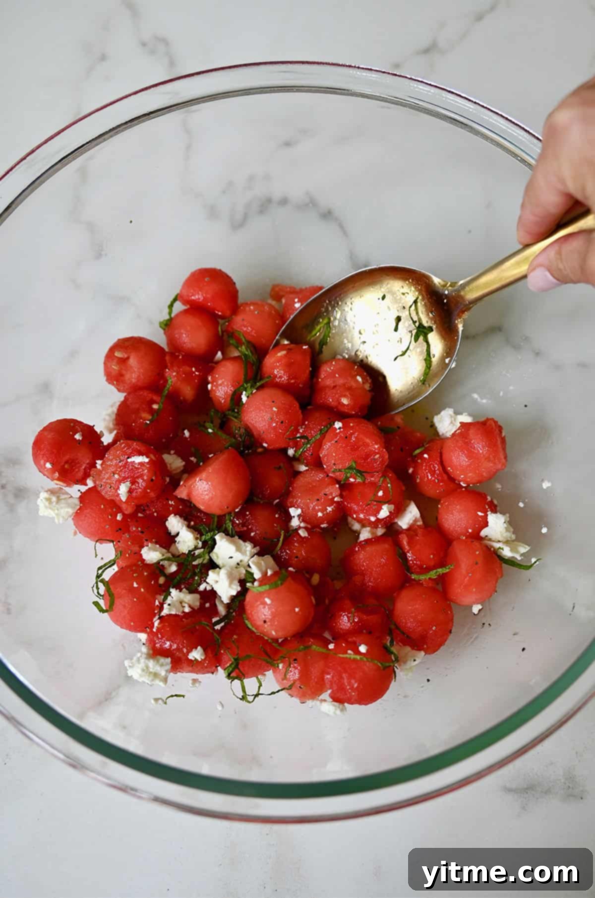 Watermelon balls with crumbled feta and fresh mint being tossed with a tangy lime dressing in a large bowl