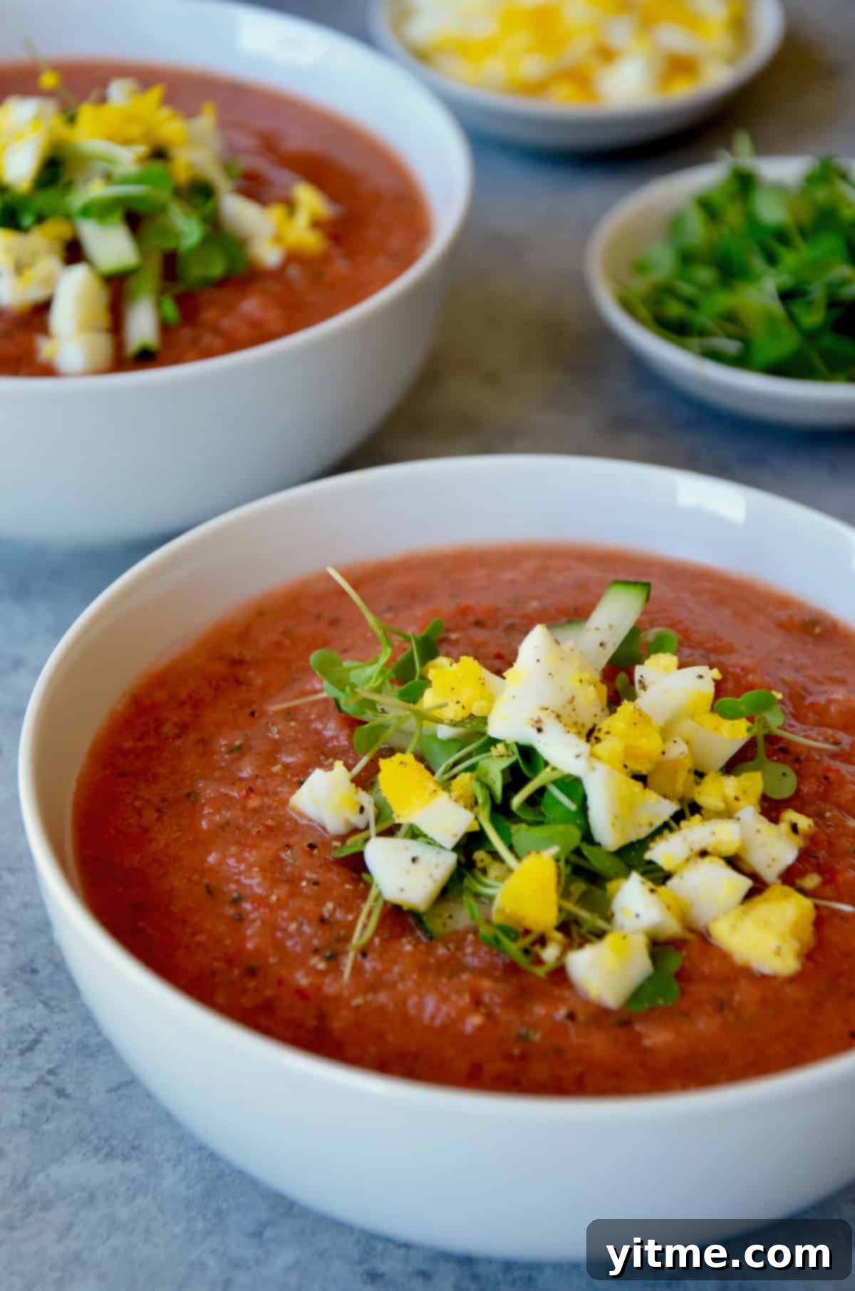 A white bowl of gazpacho garnished with chopped egg and microgreens.