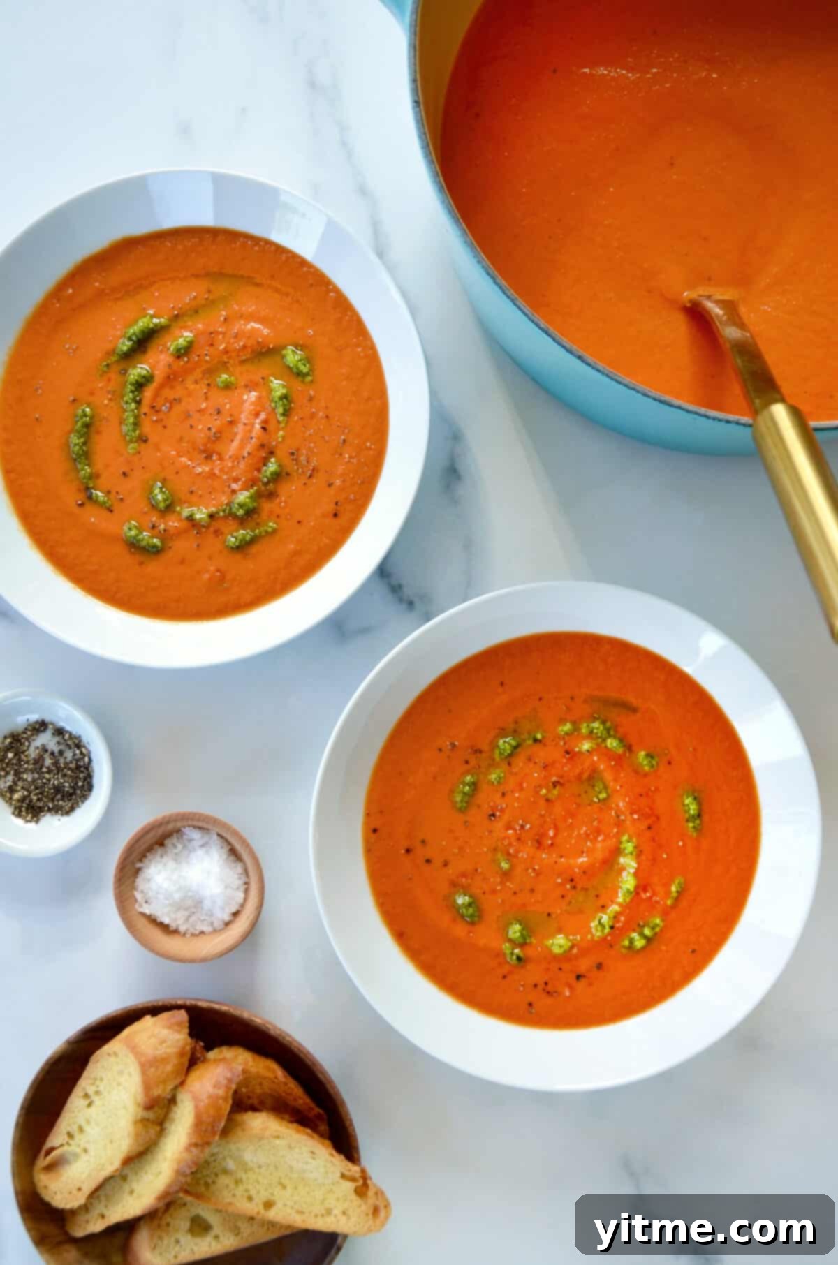 Two white bowls are filled with creamy tomato soup, elegantly drizzled with vibrant basil pesto. Nearby, small bowls hold fresh salt, cracked black pepper, and perfectly sliced baguette, inviting a delightful meal.
