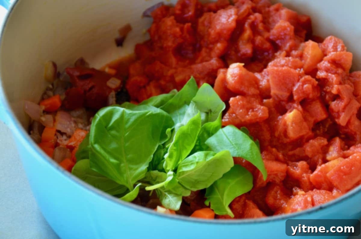 Canned San Marzano tomatoes and a sprig of fresh basil are added to a light-blue Dutch oven, symbolizing the heart of the tomato soup recipe.