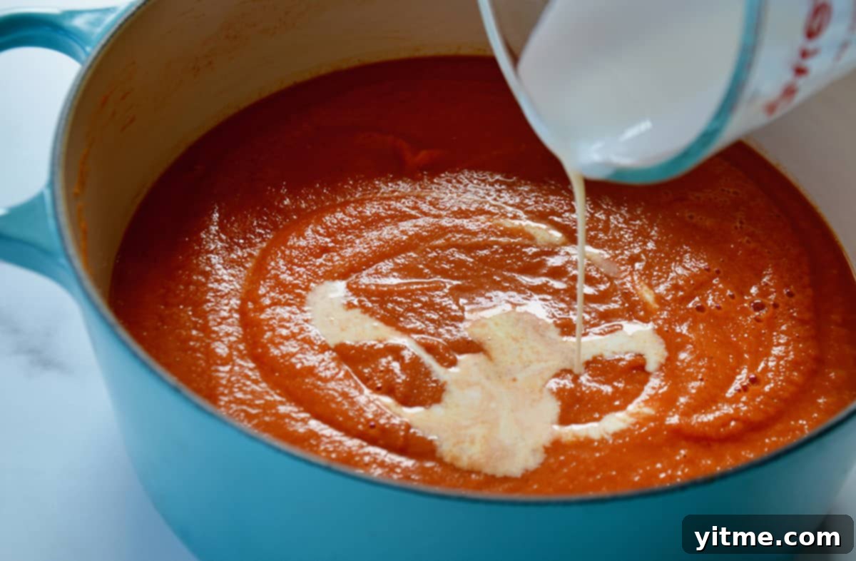 A glass measuring cup pours rich heavy cream into the vibrant tomato soup simmering in a light blue Dutch oven, signifying the final step to achieve its creamy texture.