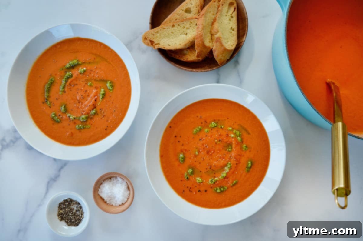 Two white bowls are generously filled with creamy tomato soup, beautifully garnished with fresh basil pesto. On the side, small dishes hold coarse salt, ground pepper, and artisan sliced baguette, inviting enjoyment.