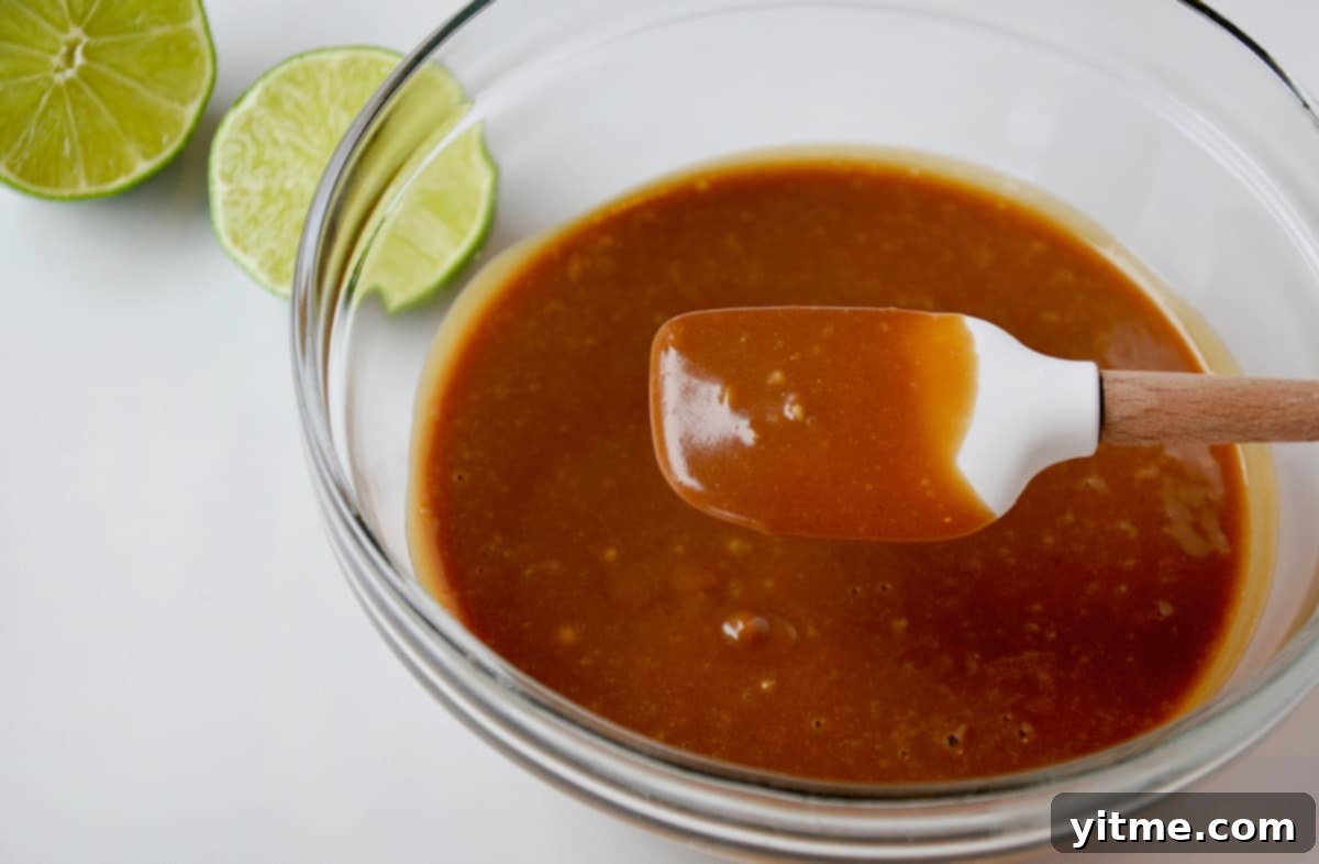A small silicone spatula in a bowl of creamy peanut sauce. A halved lime sits beside the bowl, illustrating the dressing preparation.