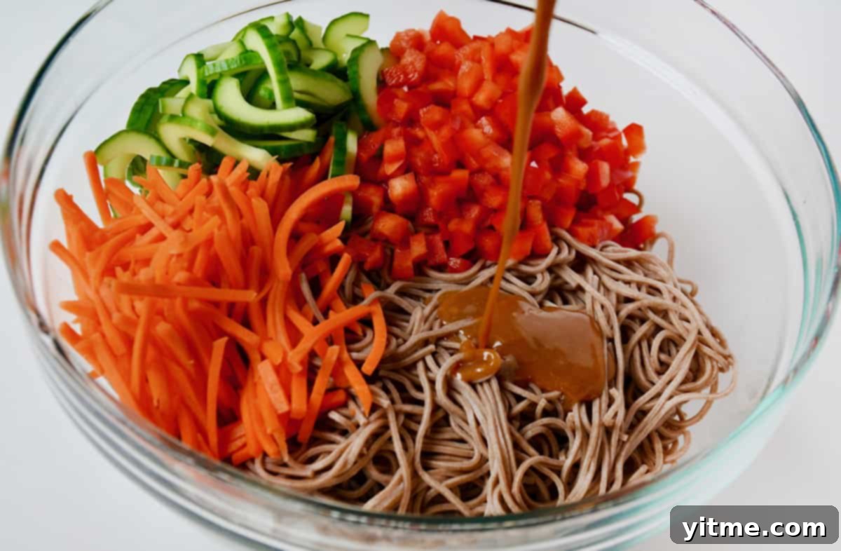 Rich peanut dressing being poured generously onto cooked soba noodles, diced red bell pepper, sliced cucumber and julienned carrots in a large glass mixing bowl, mid-preparation.