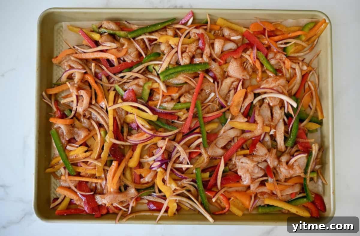 A baking sheet topped with uncooked chicken strips, sliced bell peppers and red onions all tossed in a homemade fajita seasoning.