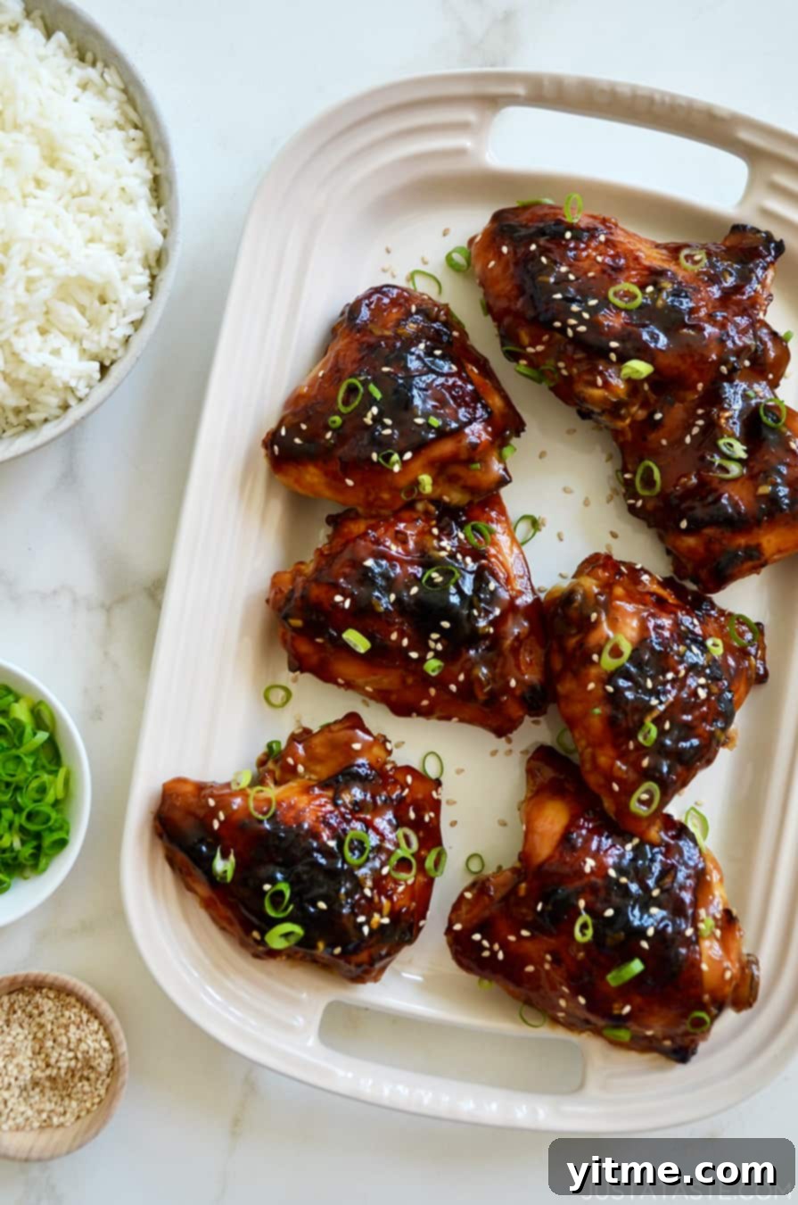 A top-down view of Glazed Honey Soy Chicken Thighs on a white serving platter next to bowls containing white rice, sliced scallions and sesame seeds