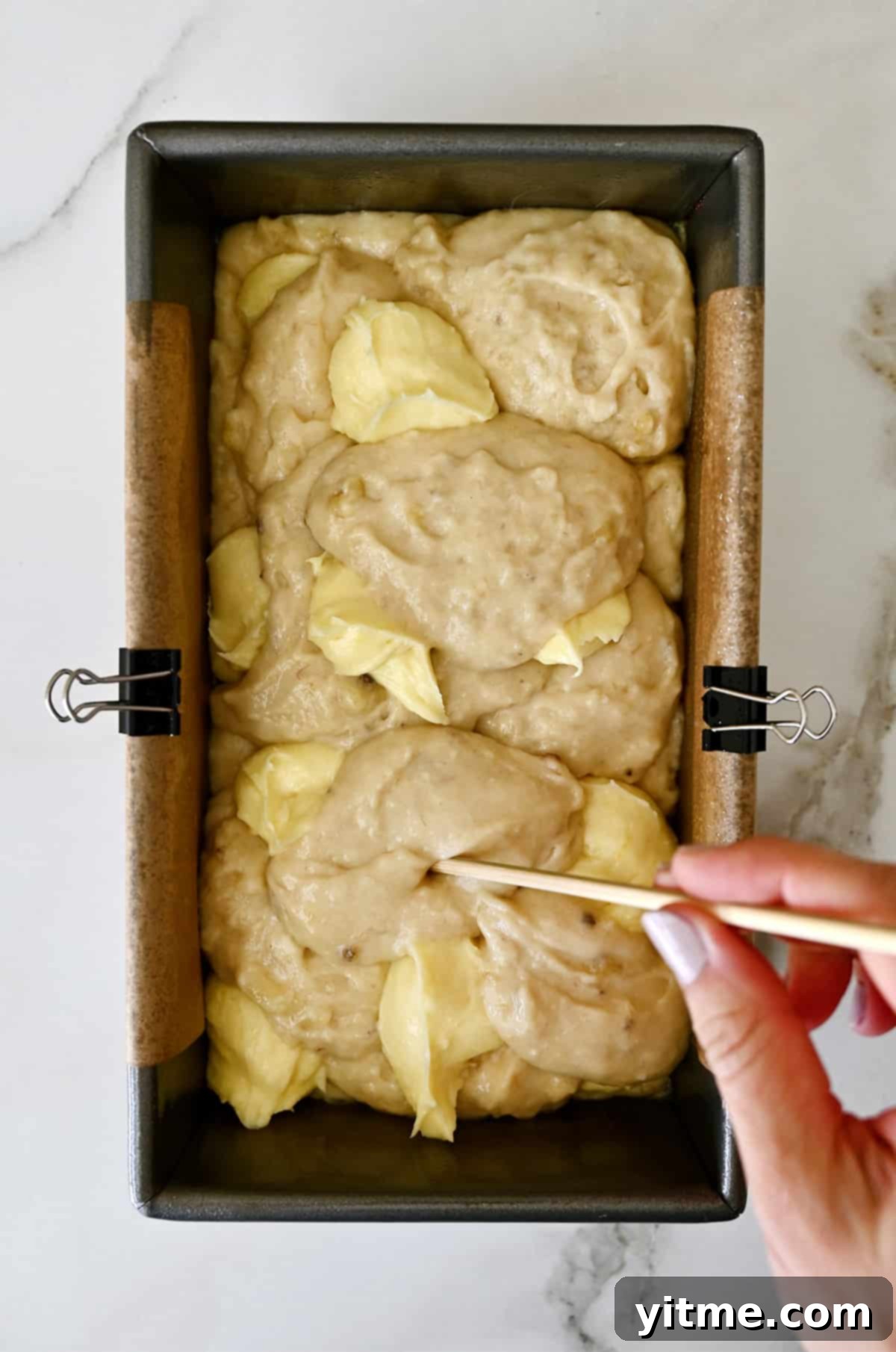 A hand using a wooden skewer to swirl cream cheese filling together with banana bread batter in a parchment paper-lined loaf pan.