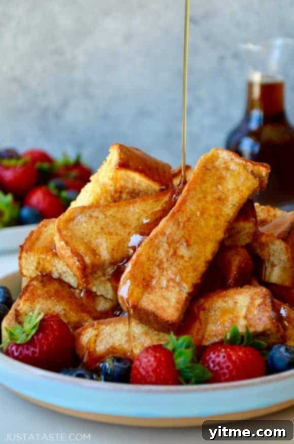 Golden baked French toast sticks arranged on a plate with fresh fruit and warm maple syrup being drizzled over the top.