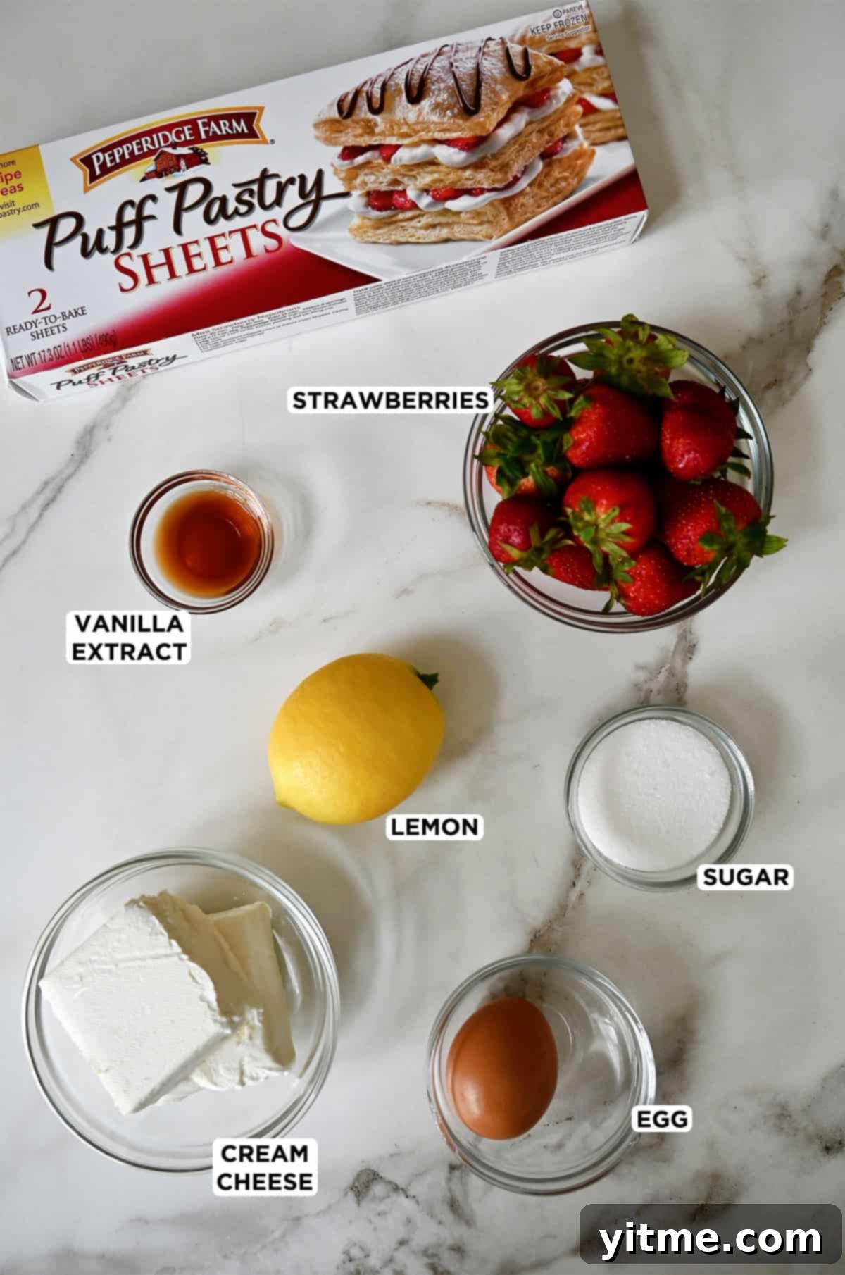 A box of puff pastry next to a lemon and various sizes of glass bowls containing vanilla extract, strawberries, cream cheese and an egg, ready for baking.