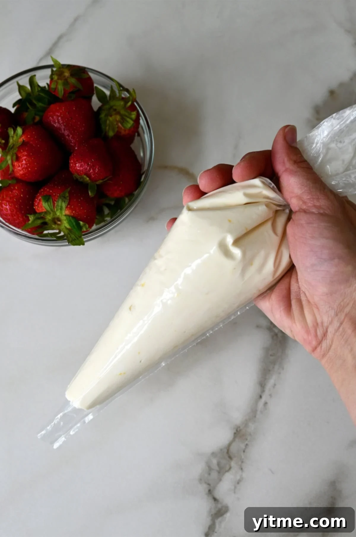 A hand holds a plastic piping bag containing lemony cream cheese filling for Danish pastries, with a bowl of fresh strawberries nearby.