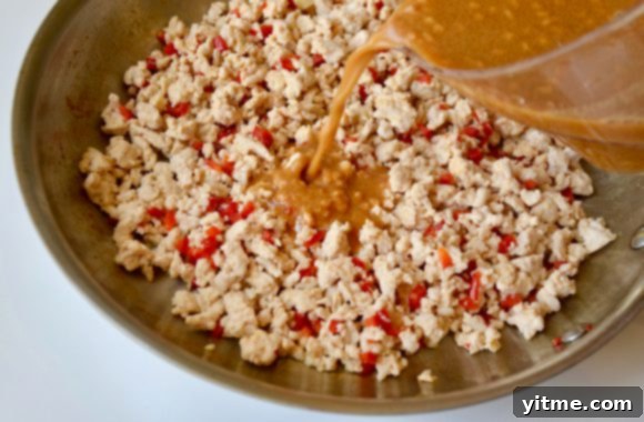 Large sauté pan containing ground turkey and diced red pepper with peanut sauce being added, showcasing the cooking process