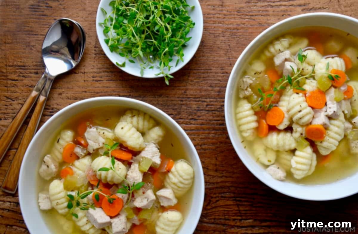 Two bowls containing homemade chicken soup with gnocchi, carrots and celery topped with a sprig of fresh thyme. Soup spoons and a bowl filled with fresh thyme is nearby.