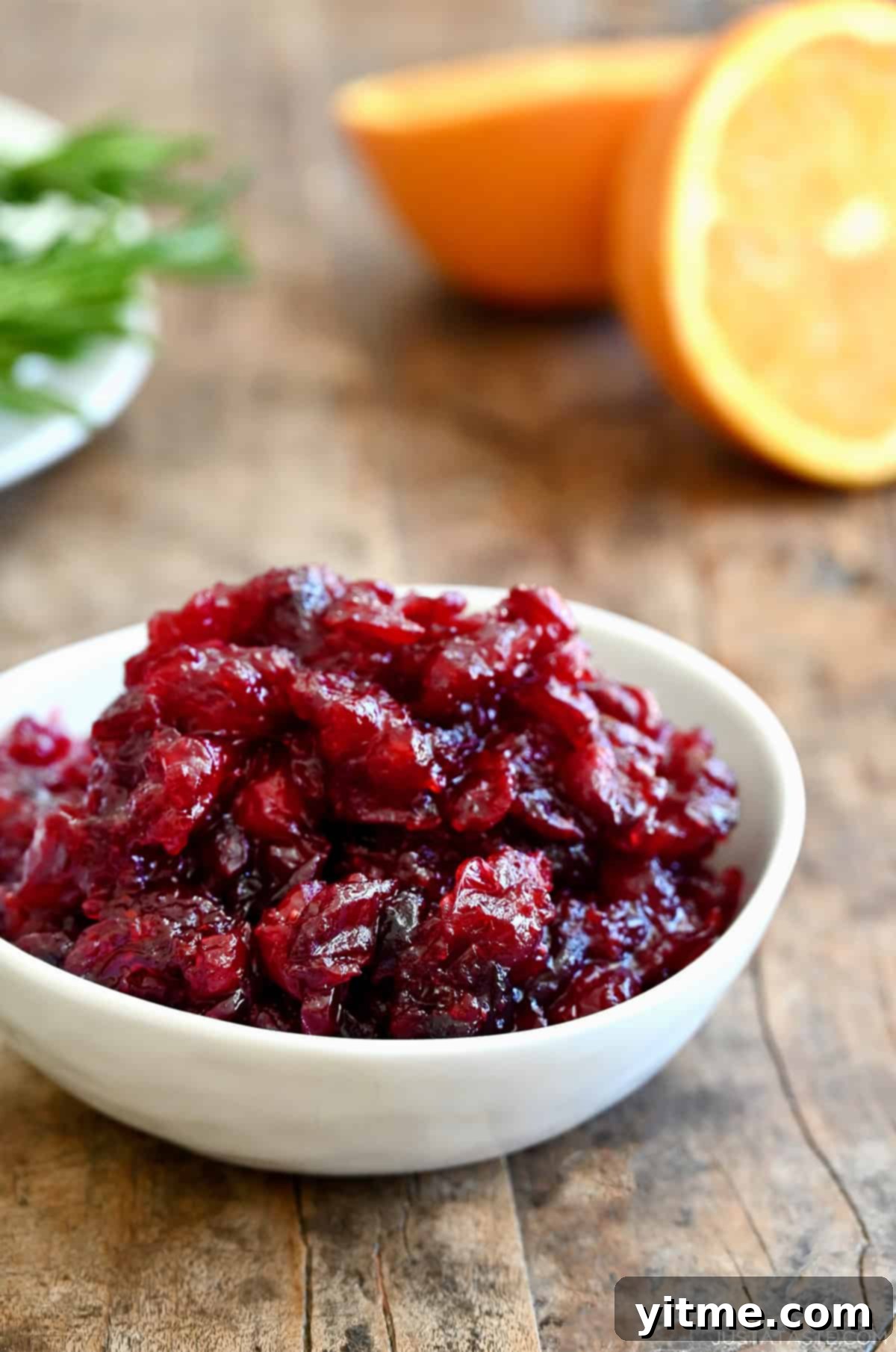 Homemade cranberry sauce in a white bowl with an orange cut in half nearby, garnished with orange zest.