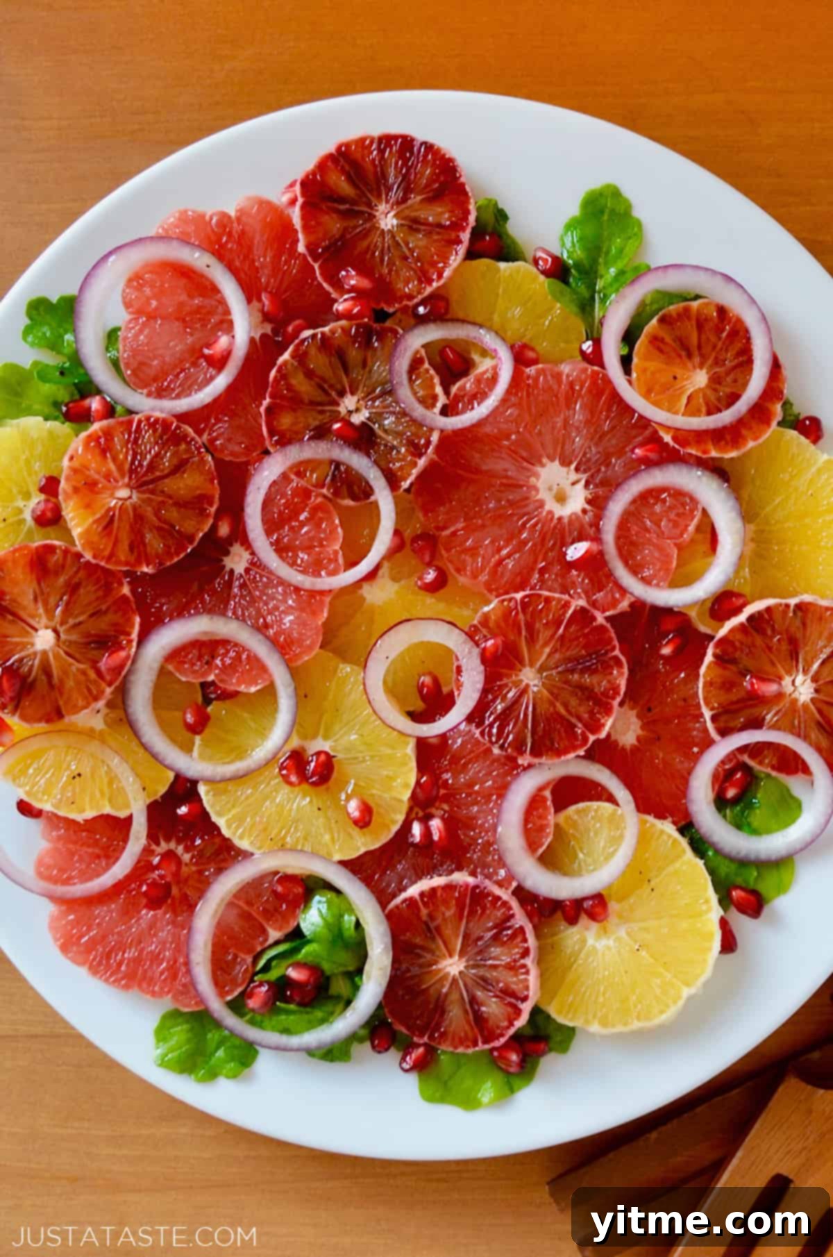 A salad comprised of sliced oranges, blood oranges and grapefruits, red onion slices, arugula and pomegranate seeds on a white serving platter. Salad tongs sit beside the platter.