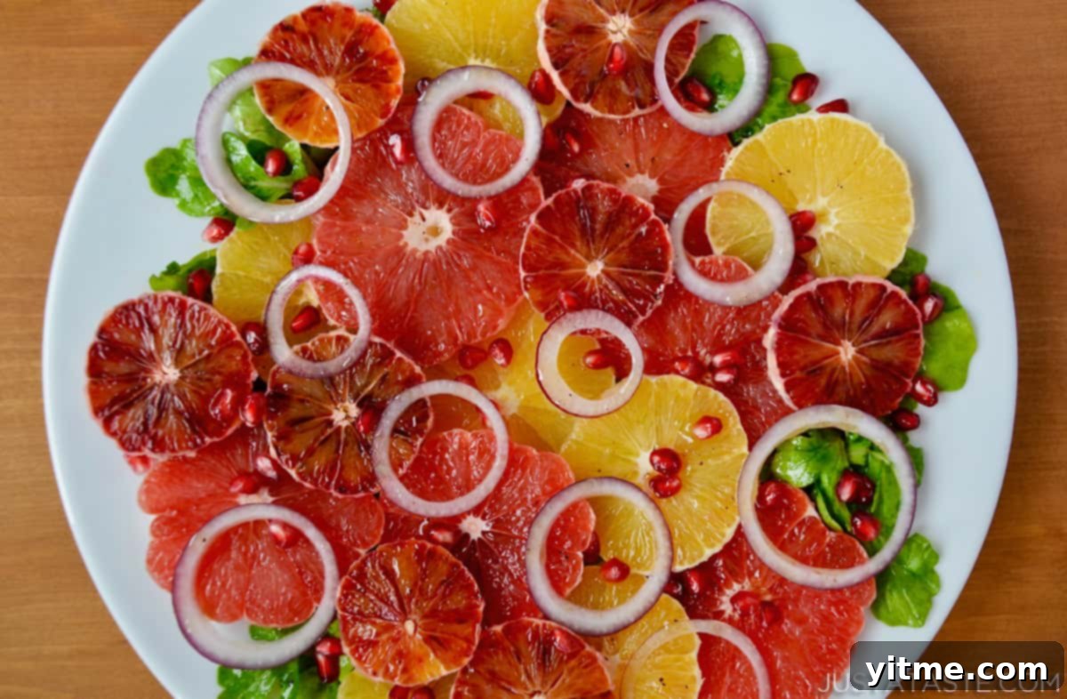 A salad of sliced oranges, blood oranges and grapefruits, red onion slices, arugula and pomegranate seeds on a white serving platter.