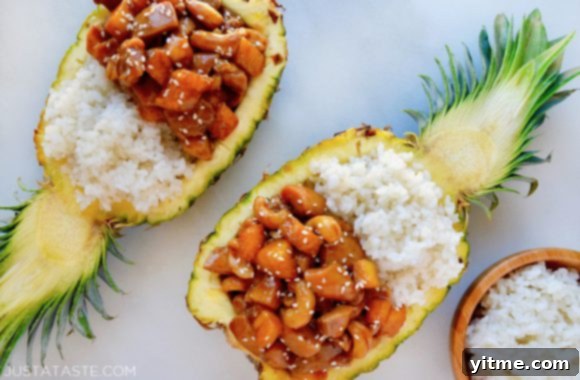 A creatively hollowed-out halved pineapple serves as a bowl, filled with fluffy white rice and generously topped with glistening Sticky Pineapple Chicken.