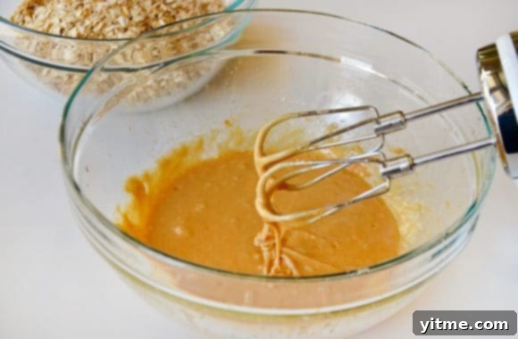 A large clear glass bowl filled with creamy peanut butter, golden honey, a fresh egg, clear oil, and aromatic vanilla extract, ready for mixing with a handheld beater.