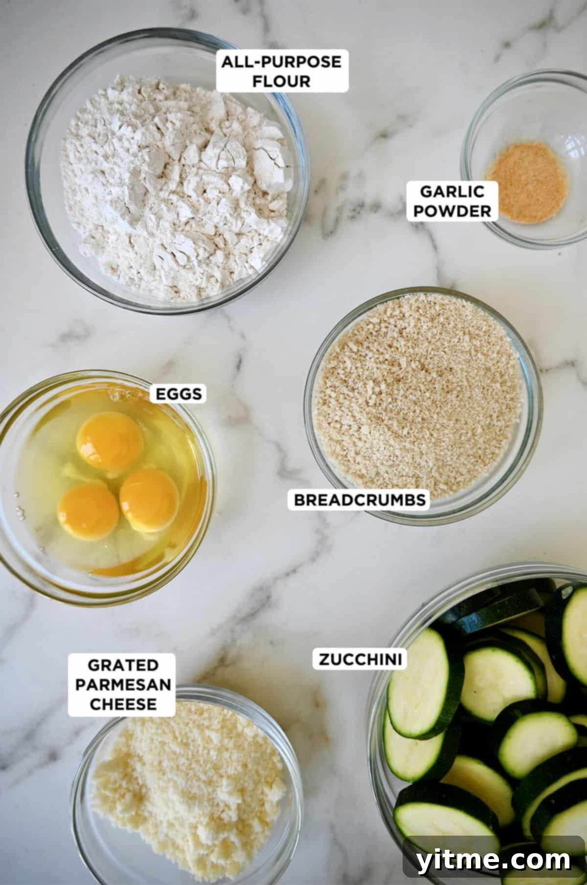 Ingredients to make zucchini chips in glass bowls, including sliced zucchini, grated Parmesan cheese, eggs, breadcrumbs, flour and garlic powder.