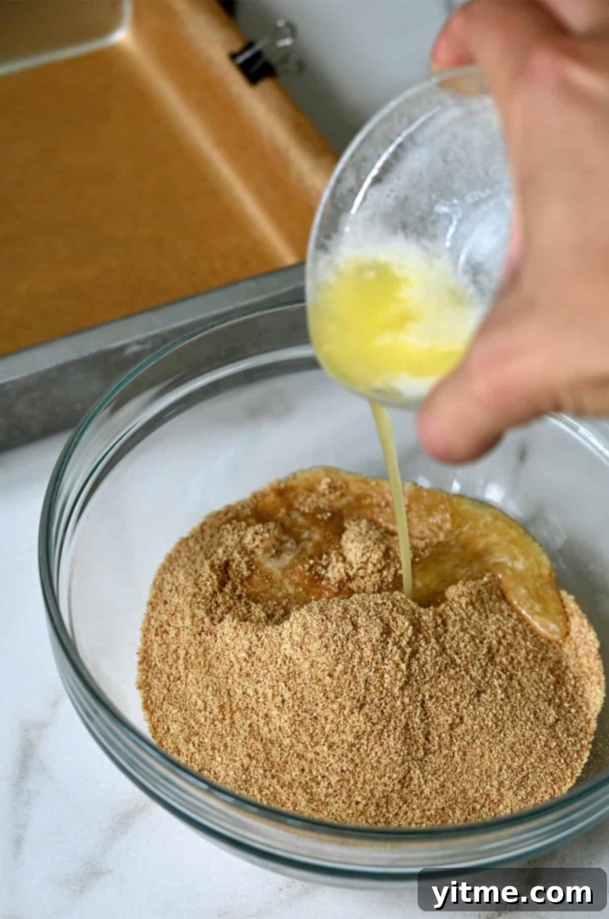 Melted butter being poured from a small bowl atop graham cracker crumbs in a bowl.