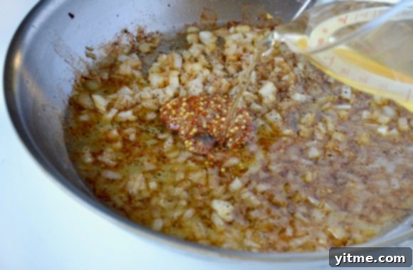 Large sauté pan with diced onions and whole grain mustard with apple cider vinegar being poured from glass measuring cup.