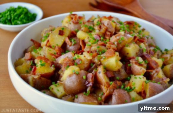 White bowl with Potato Salad with Warm Bacon Dressing garnished with fresh chives with small white bowl filled with fresh chives and wooden serving utensils in background.