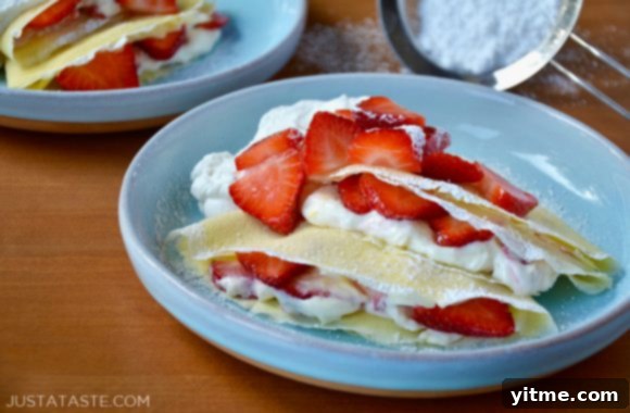 Strawberry Dream Crêpes 6 Strawberry Cream Cheese Crepes with fresh strawberries, whipped cream, and powdered sugar, with a mesh sifter in the background.