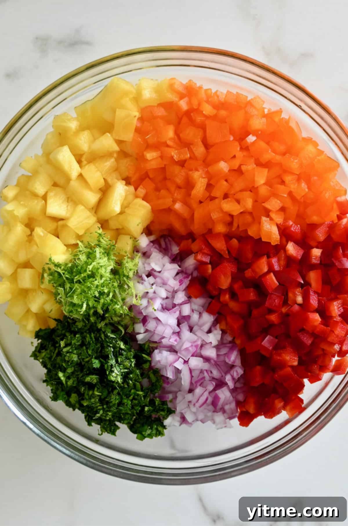 A clear bowl showcasing the vibrant chopped ingredients of pineapple salsa: orange and red bell peppers, diced red onion, fresh cilantro, lime zest, and small pieces of pineapple.