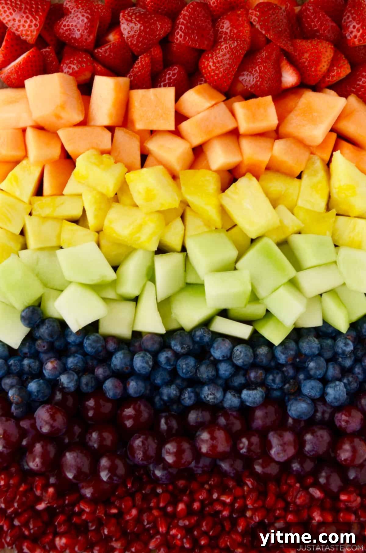 A stunning array of fresh, colorful fruits ready for preparation, including brightly sliced strawberries, sweet cantaloupe, golden pineapple, light green honeydew melon, deep blue blueberries, rich purple grapes, and glistening red pomegranate arils.