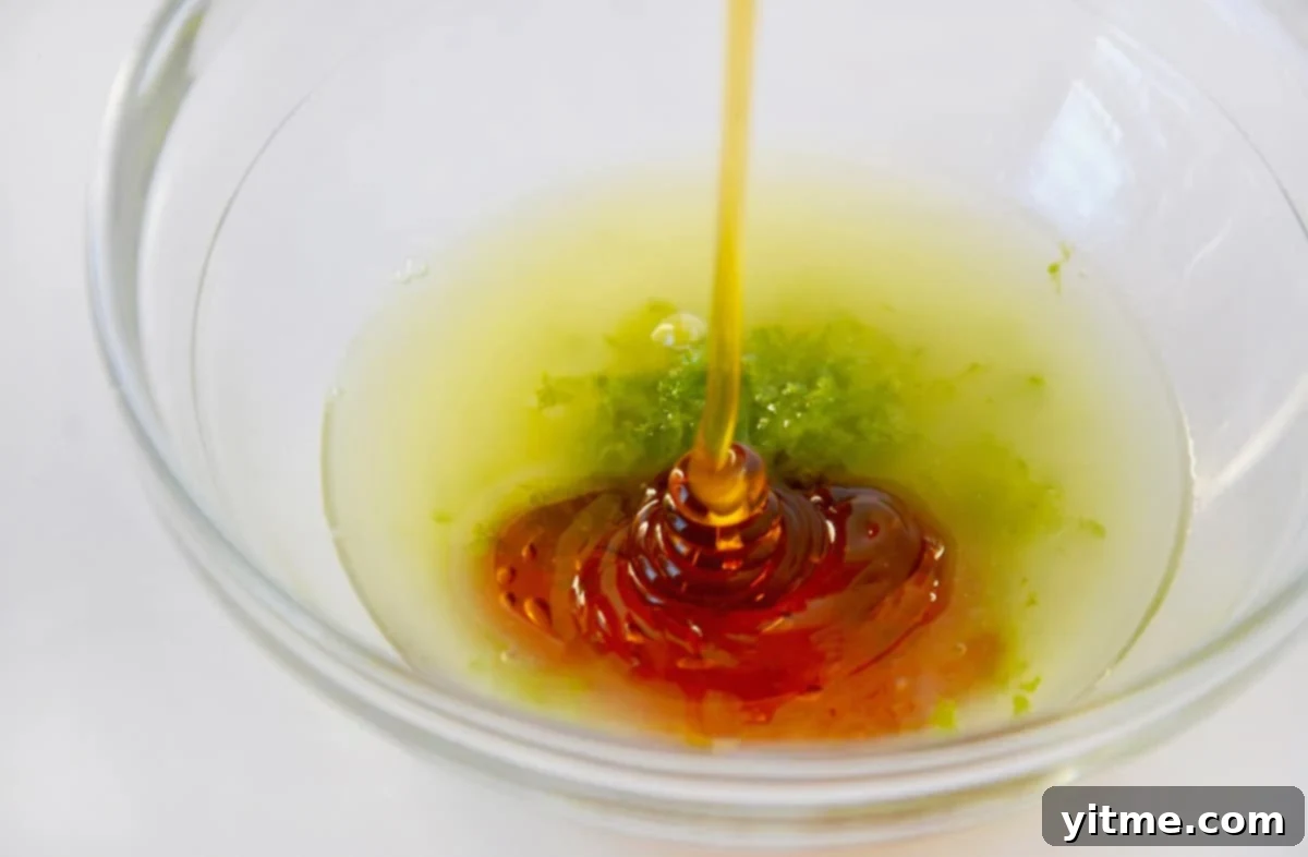 Close-up shot of golden honey being poured into a clear glass bowl containing bright green lime juice and aromatic lime zest, ready to be whisked into a refreshing fruit salad dressing.