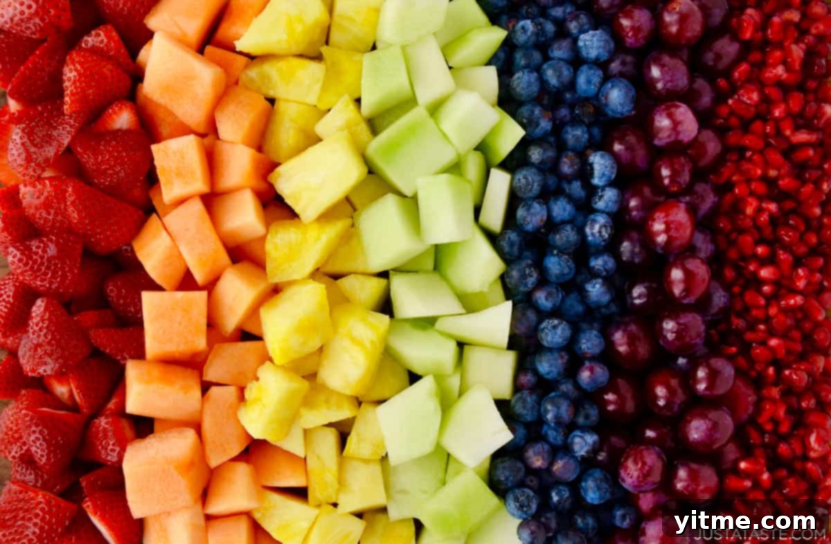 A beautifully arranged serving bowl showcasing a rainbow of fresh fruit salad, featuring sliced strawberries, golden cantaloupe, sweet pineapple, light green honeydew melon, deep blue blueberries, juicy purple grapes, and sparkling pomegranate seeds, all tossed with a light dressing.
