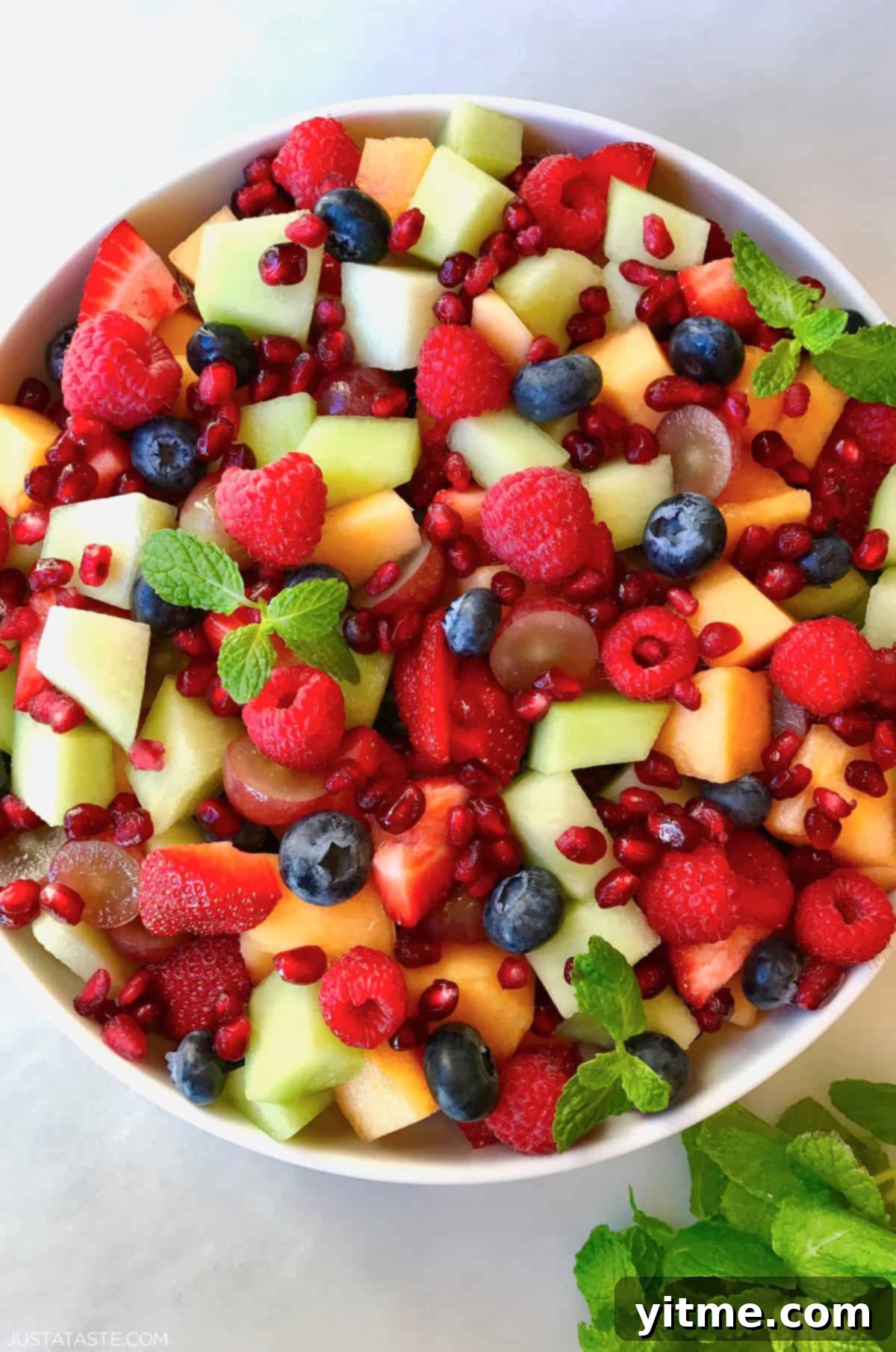 A large serving bowl containing a refreshing fruit salad with diced cantaloupe and honeydew melon, blueberries, raspberries and pomegranate arils, drizzled with honey-lime dressing.