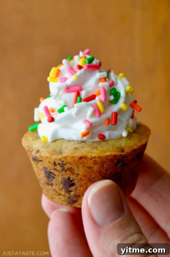 Muffin Tin Chocolate Chip Cookie Treats 3 A close-up shot of a hand holding a Chocolate Chip Cookie Cup topped with vanilla frosting and sprinkles