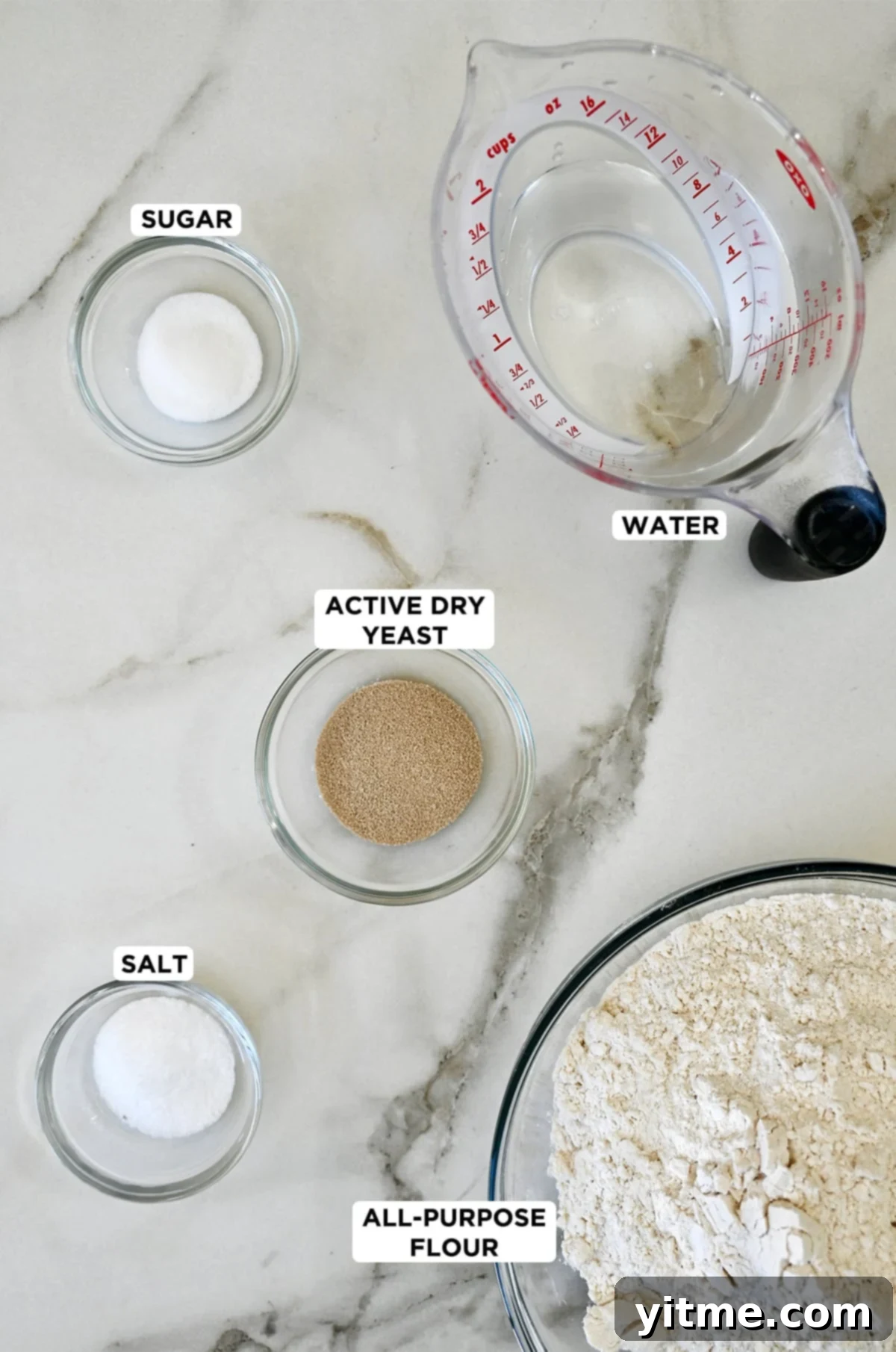 Various essential pizza dough ingredients are neatly arranged in glass bowls: sugar, active dry yeast, kosher salt, and all-purpose flour. Beside them, a liquid measuring cup holds the perfect amount of water.