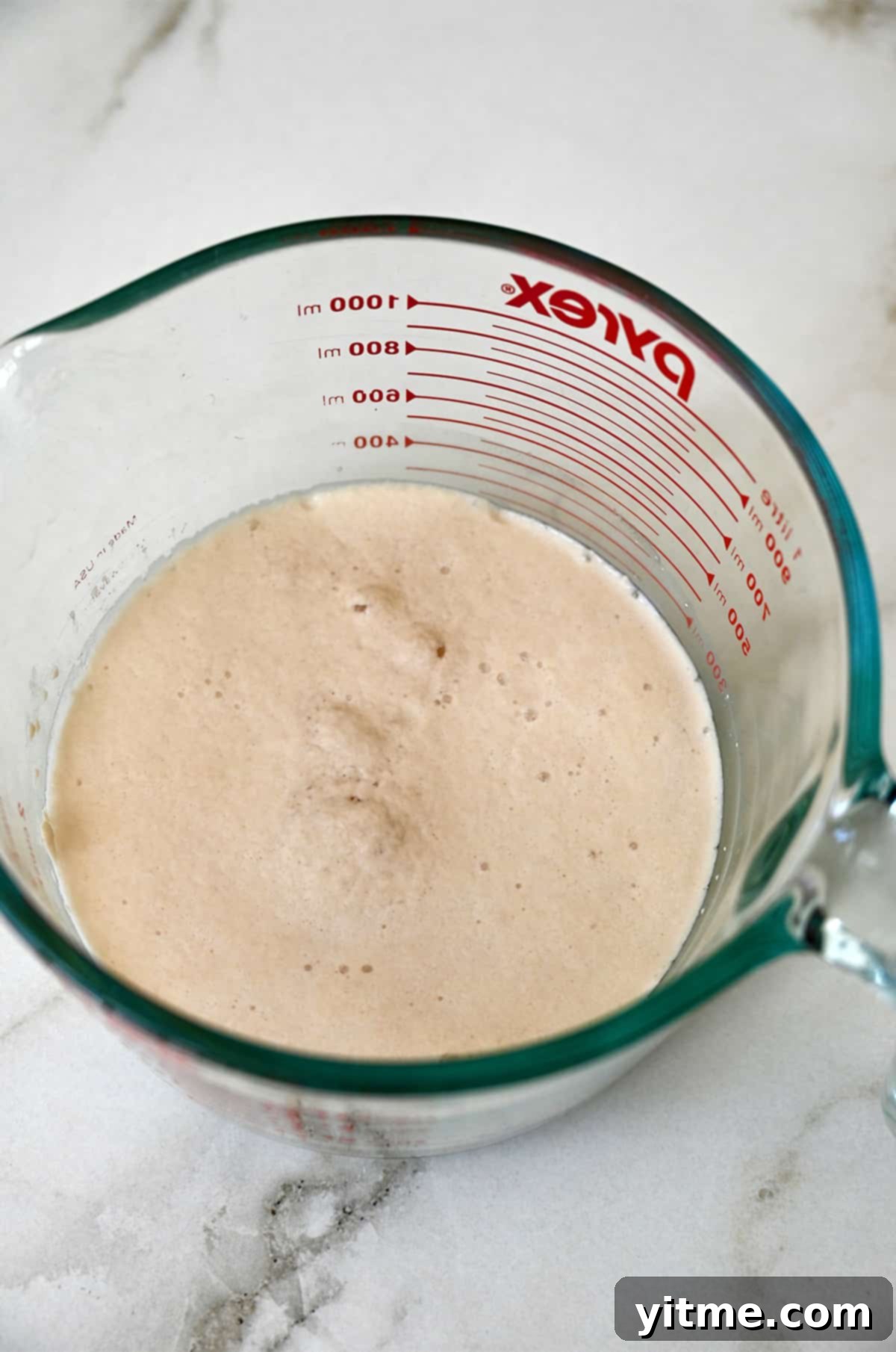 A frothy mixture of activated yeast, sugar, and warm water is shown in a clear liquid measuring cup, ready for the next step of dough preparation.
