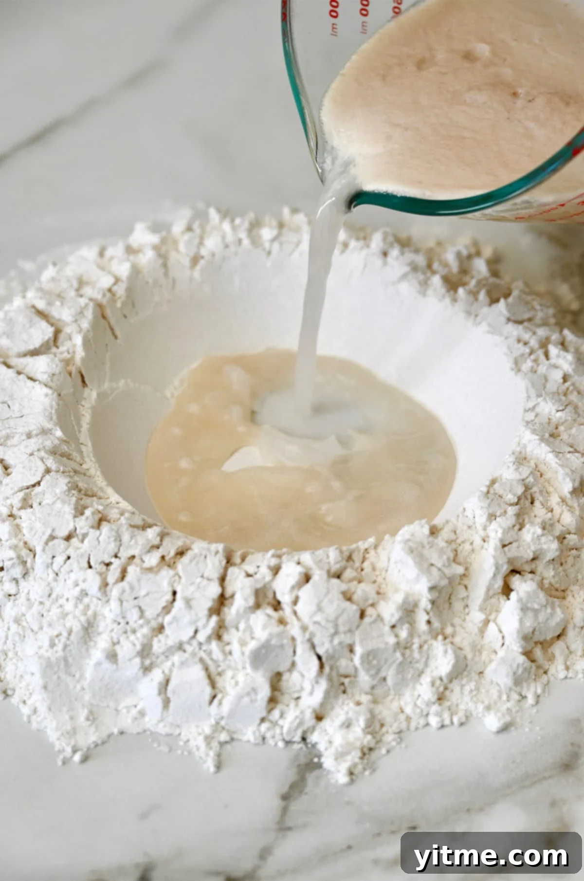 The activated yeast and water mixture is being carefully poured into a well made in a mound of all-purpose flour on a clean work surface, illustrating the start of dough formation.
