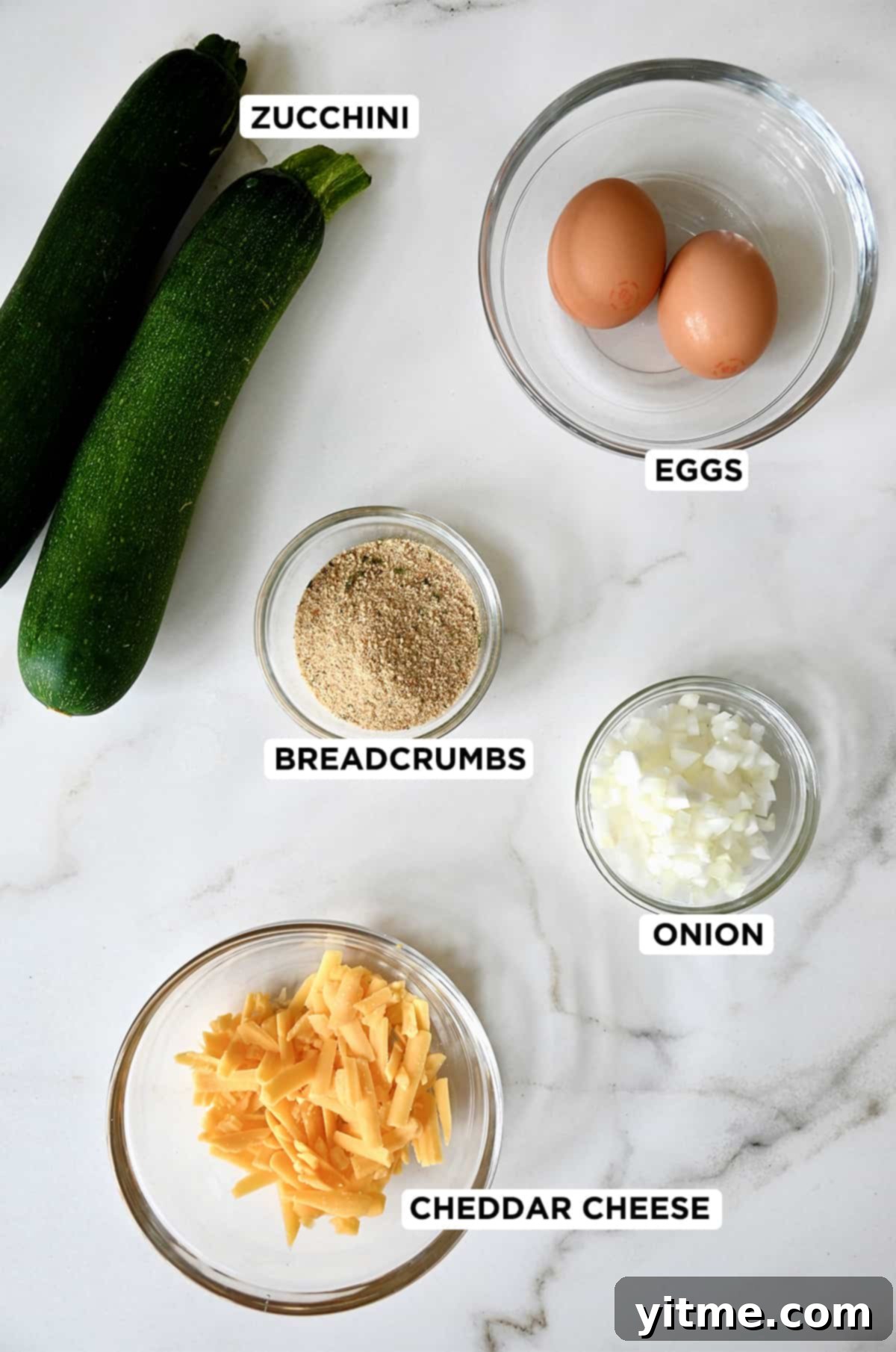 Two zucchinis next to various sized of clear bowls containing eggs, diced onion, breadcrumbs and shredded cheddar cheese.