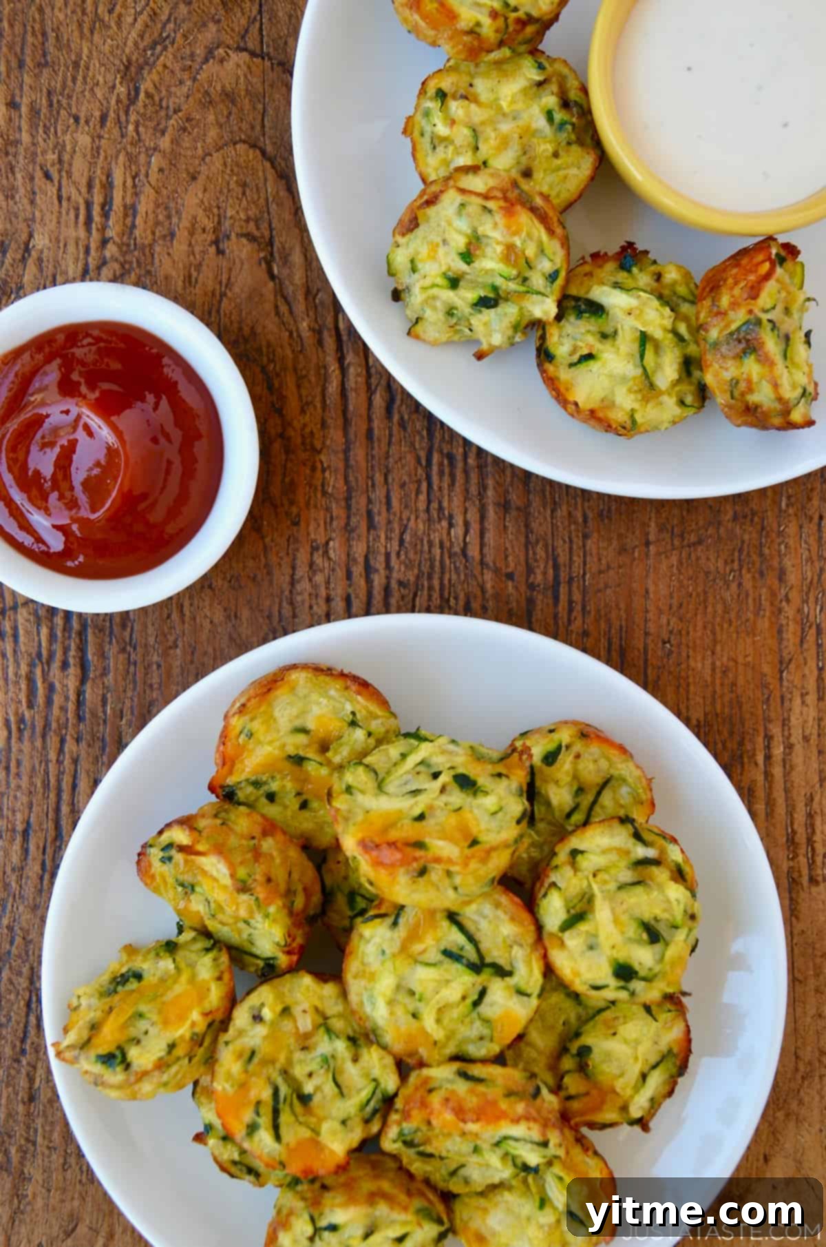 Two white dinner plates piled high with cheddar cheese zucchini tots next to a small bowl filled with ketchup and a small bowl containing ranch dressing.