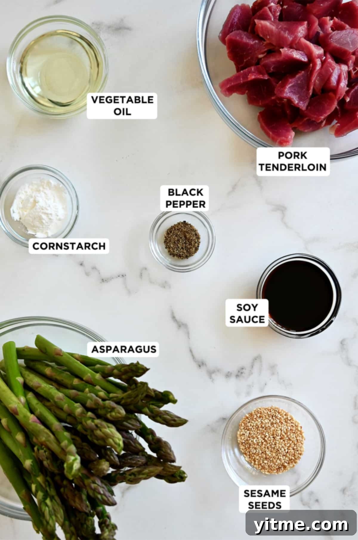Various sizes of glass bowls containing pork tenderloin, vegetable oil, black pepper, cornstarch, soy sauce, asparagus and sesame seeds, ready for cooking.