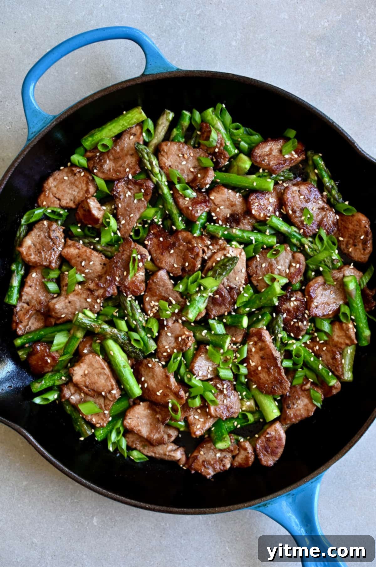 Black pepper pork and asparagus stir-fry in a skillet, garnished with toasted sesame seeds and sliced scallions, ready for serving.
