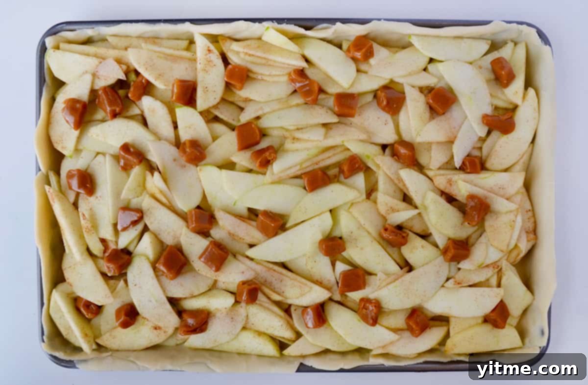 A sheet pan containing pie dough topped with apple slices, soft caramels and ground cinnamon.