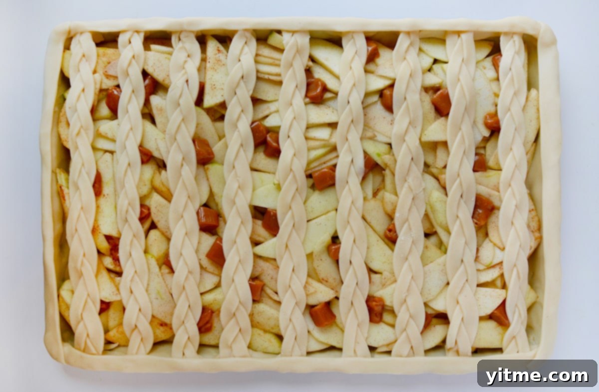 A sheet pan with a fully assembled unbaked pie with a bottom crust, apple filling, soft caramels and braided lattice top crust.