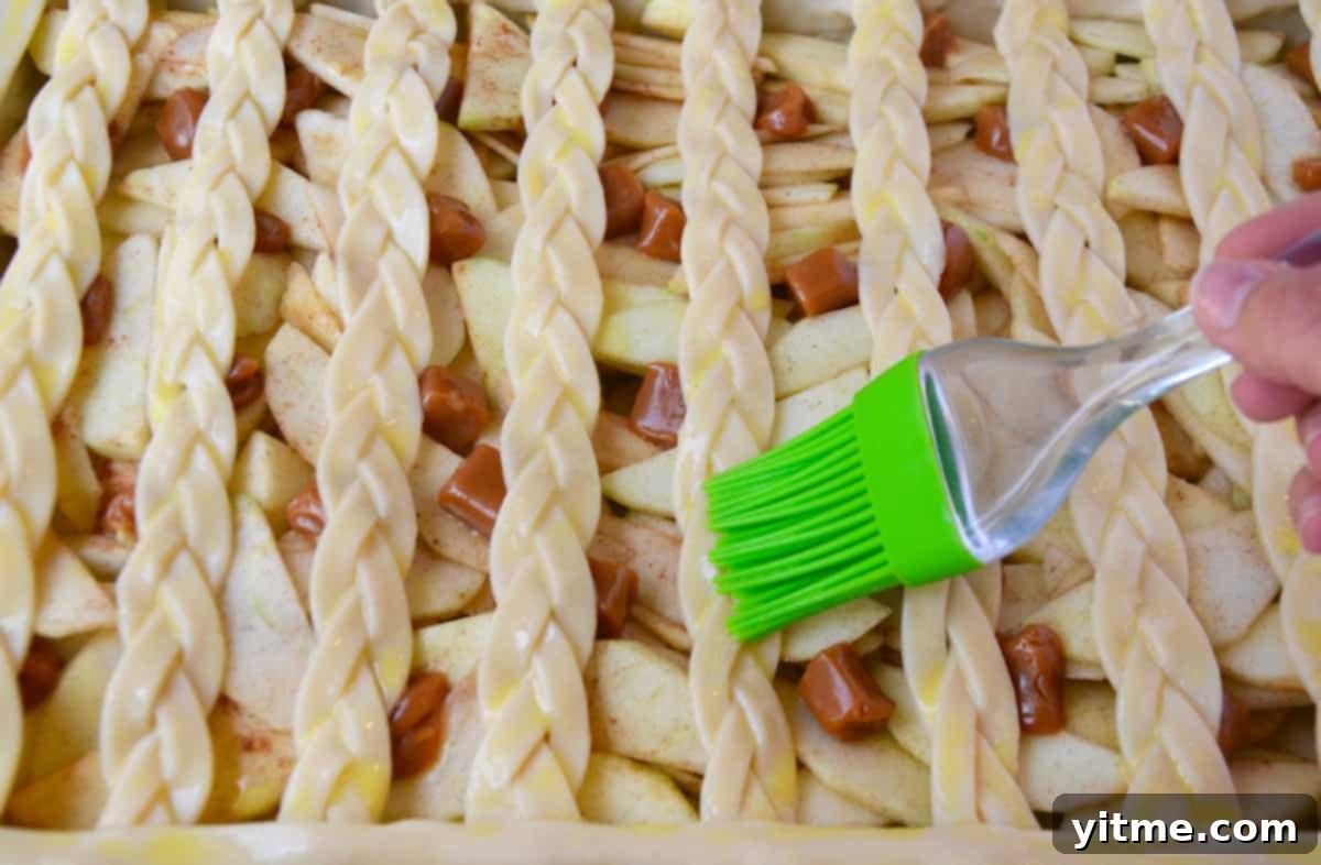 A hand using a pastry brush to brush egg wash onto the braided lattice pie crust before baking.