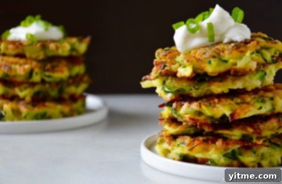 Two white plates showcasing stacks of golden-brown zucchini fritters, each topped with a dollop of creamy sour cream. A simple and impressive dish that has become a Just a Taste classic.