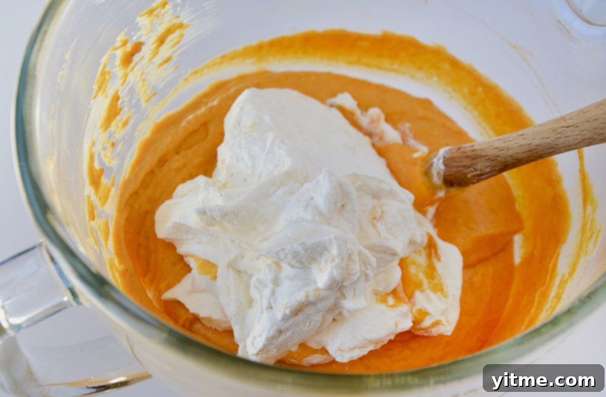 Whipped cream on top of pumpkin filling in the bowl of a stand mixer. A spatula is propped up against the side of the bowl.