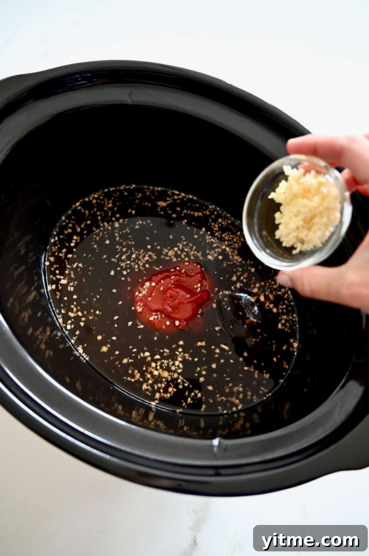 Adding minced garlic and ginger to a slow cooker with soy sauce, chicken stock, pineapple juice, ketchup, and brown sugar