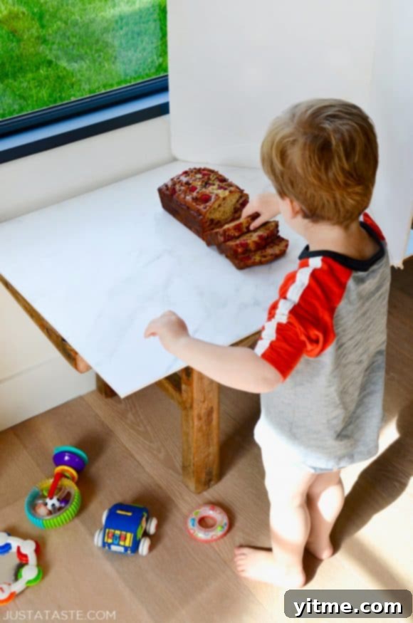 A child stands proudly in front of a freshly baked loaf of Raspberry Chocolate Chip Banana Bread, highlighting the recipe's kid-friendly appeal.